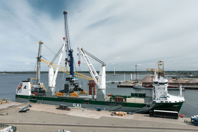 A green and white heavy-lift cargo ship moored at a quay under a cloudy sky. Its white deck cranes are lifting crane components aboard, while a tall blue lattice crane boom rises above the ship. Wind turbines and other port facilities are visible across the water in the background.