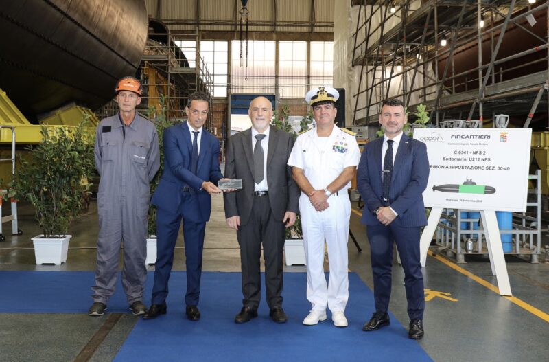 Guests, including an Italian Navy officer and a shipyard worker, pose with a commemorative plaque at the U212NFS keel-laying ceremony at Fincantieri's Muggiano shipyard