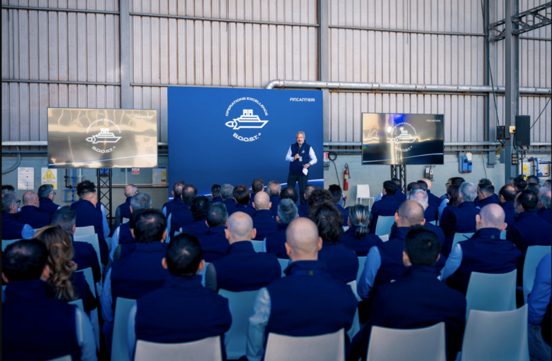 Speaker addressing Fincantieri employees in front of an Operations Excellence BOOST² backdrop at a presentation inside a shipyard building